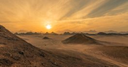 A photo of a sunset in the desert. It is a flat sandy lanscape, with several small hills popping up here and there. You can just make out two people who have climbed up a mountain in the distance. The location is Qesm Safaga, Red Sea Governorate, Egypt.